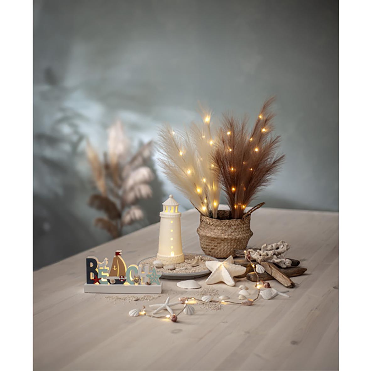 Dekorationskvist med ljusbeige plumes och små ljusslingor på ett bord med juldekorationer och en liten fyr