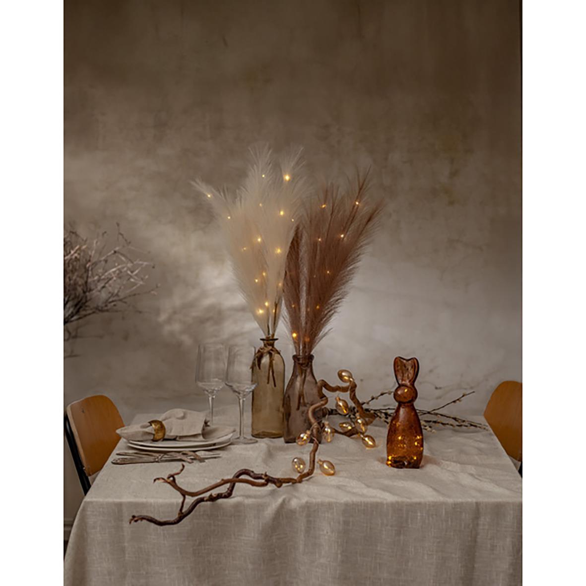 Dekorationskvist med ljusbeige plumes och små ljusdetaljer på ett dukat bord med ljus och dekorativa figurer