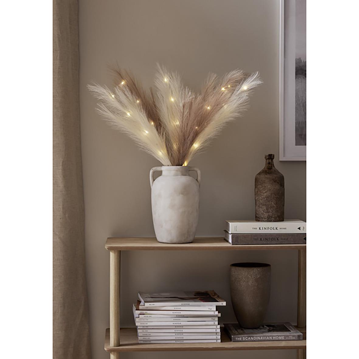 Dekorationskvist med bruna fjädrar och små ljus i ett vitt krus på ett trähyllor