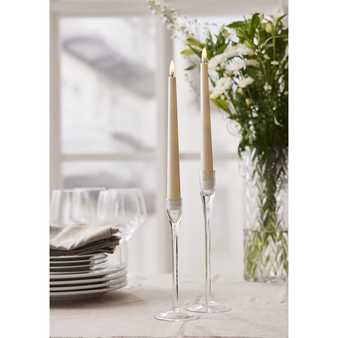 Två antika ljus i gult med glasfötter på ett bord, omgivna av tallrikar och en blomsterbukett