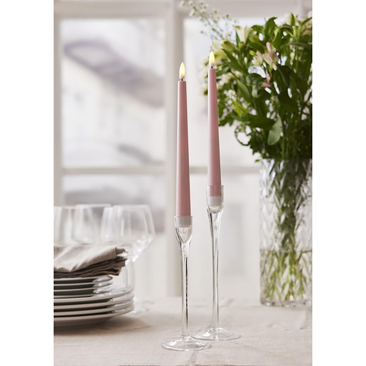 Två rosa pastellfärgade antikljus i glasljusstakar på ett bord med blommor i bakgrunden