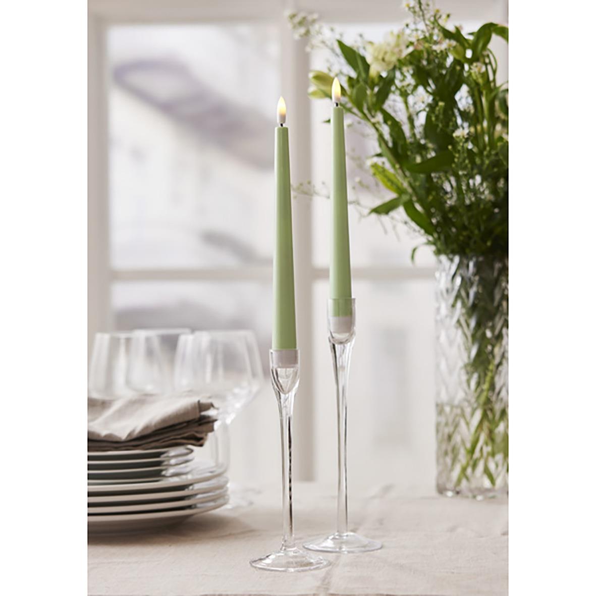 Två gröna antikljus i glasstakar på ett bord med vita tallrikar och en blomsterbukett i bakgrunden