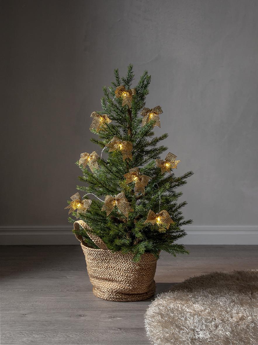 Liten julgran i kruka med guldglittrande ljusslinga och guldiga dekorationer framför en grå vägg