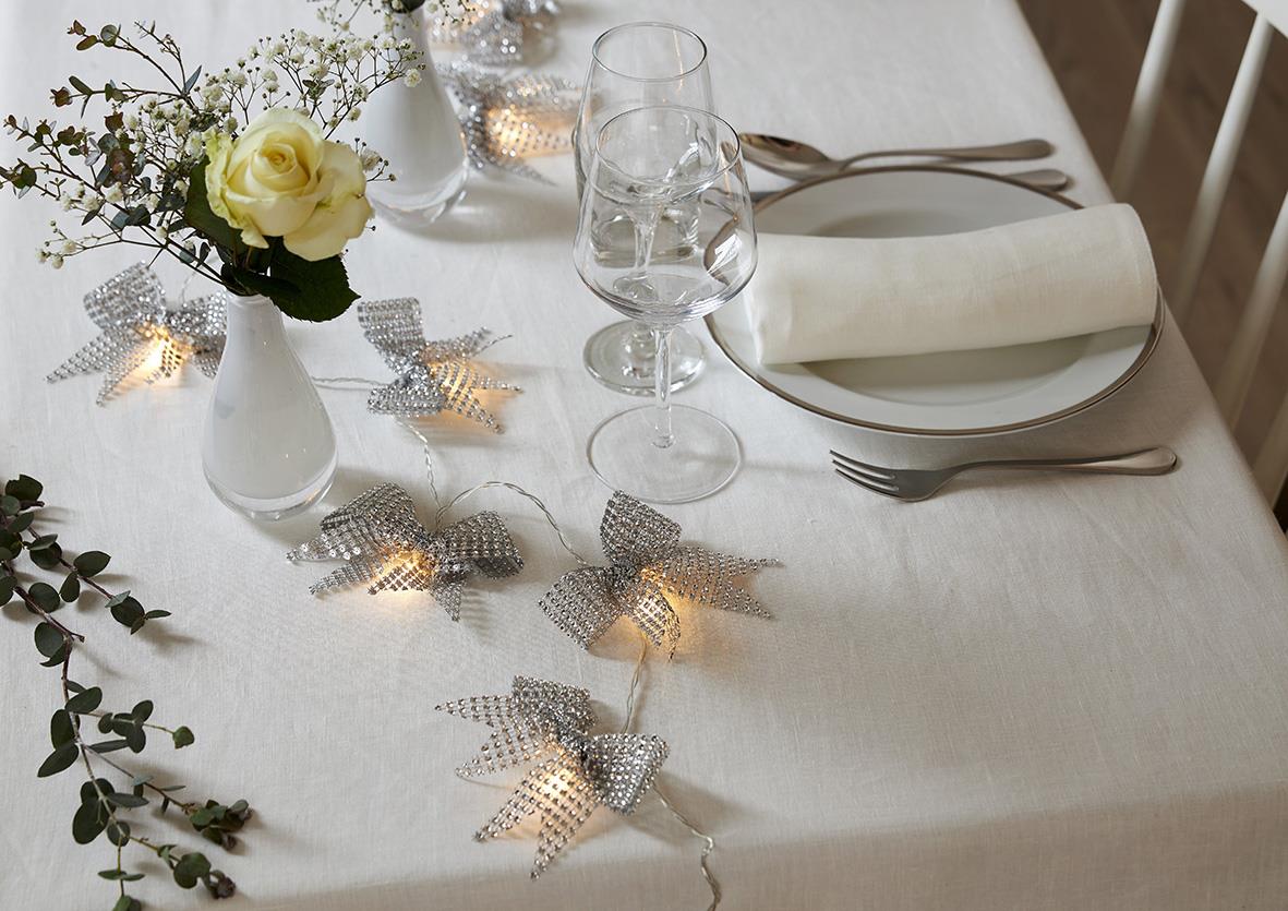 Vitt bordduk med ljusslinga Star Trading Bowie Silver, blomsterarrangemang och dukade glas och bestick