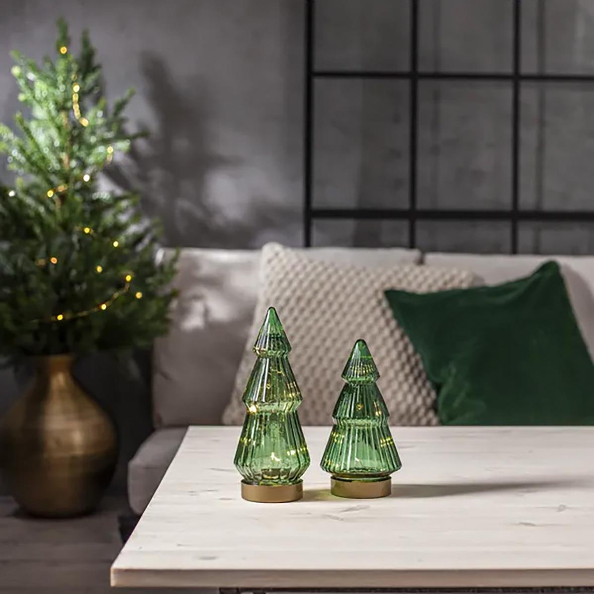 Tre gröna glasdekorationer i form av julgranar på ett vitt bord, med julgran och soffkuddar i bakgrunden