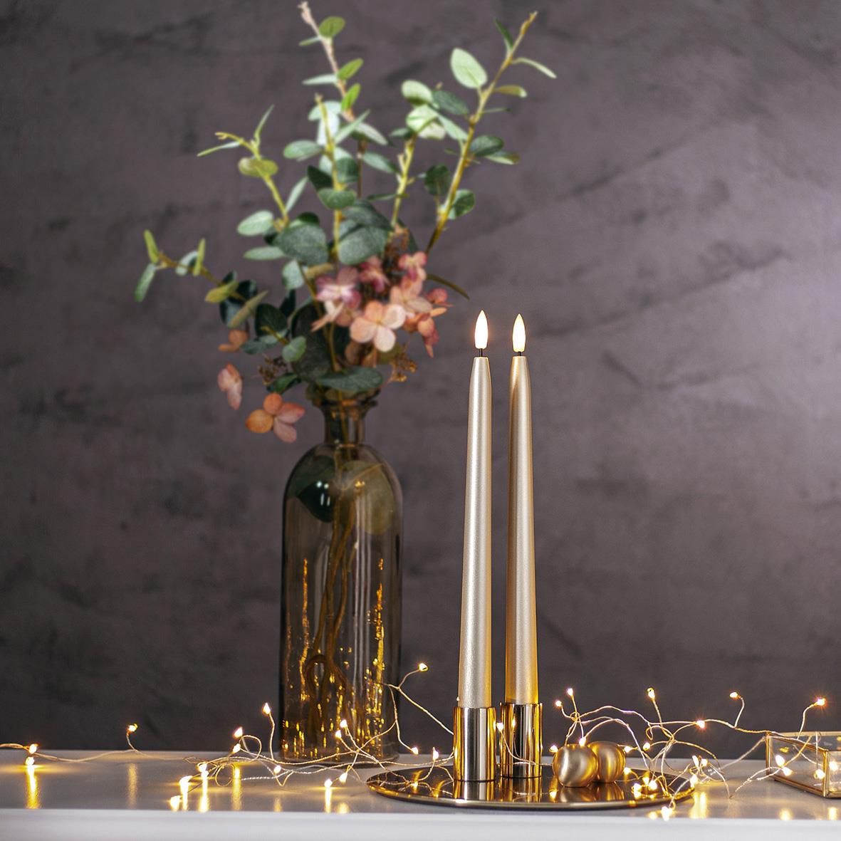 Antikljus Star Trading LED Flamme i guldfärgad metall, 28,5 cm, placerad framför en vas med gröna blad och blommor, omgiven av små ljusslingor