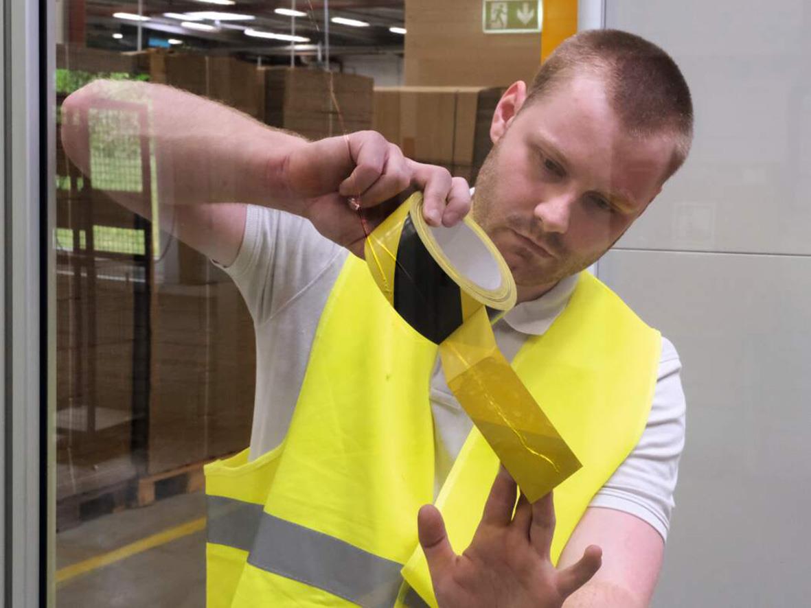 Man i reflexväst applicerar gul varningstejp på glasruta