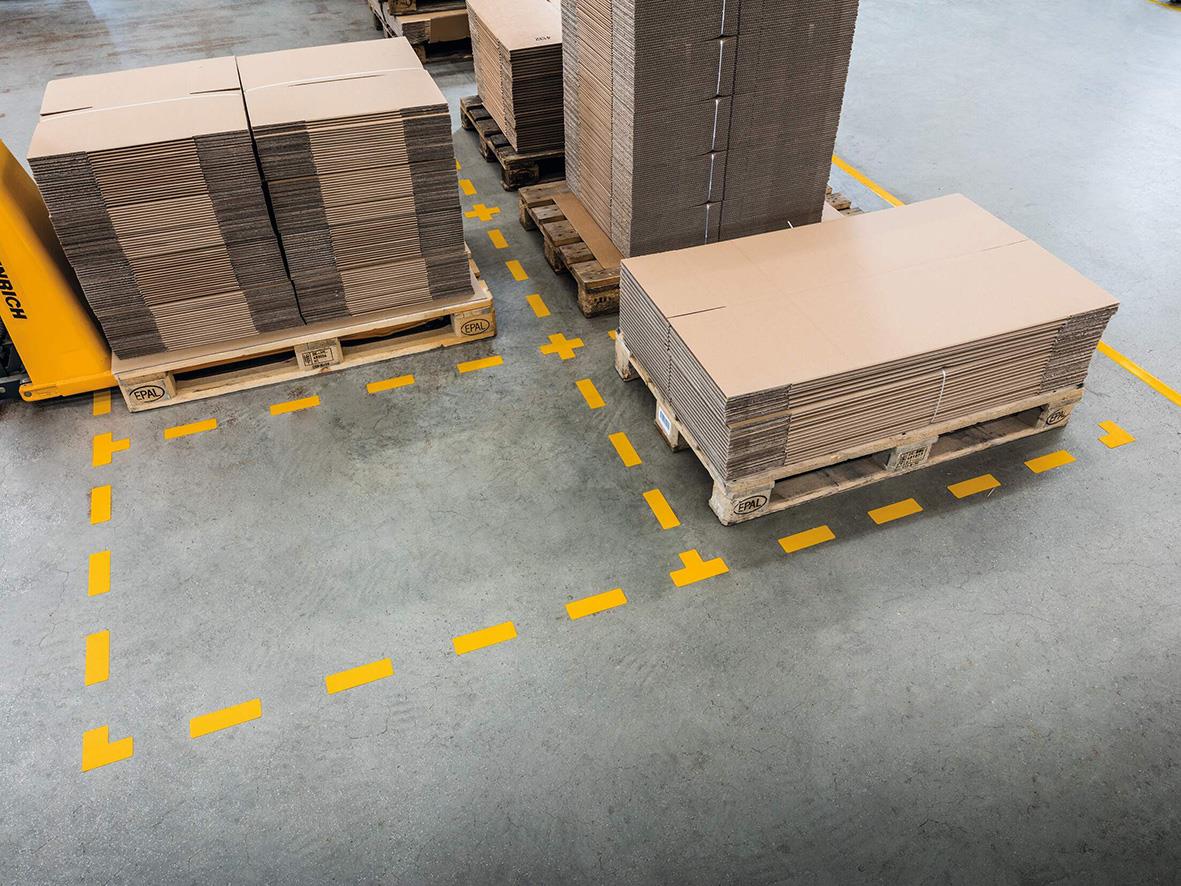 Golvmarkering i gul färg i L-form på fabriksgolv med pallar av byggmaterial