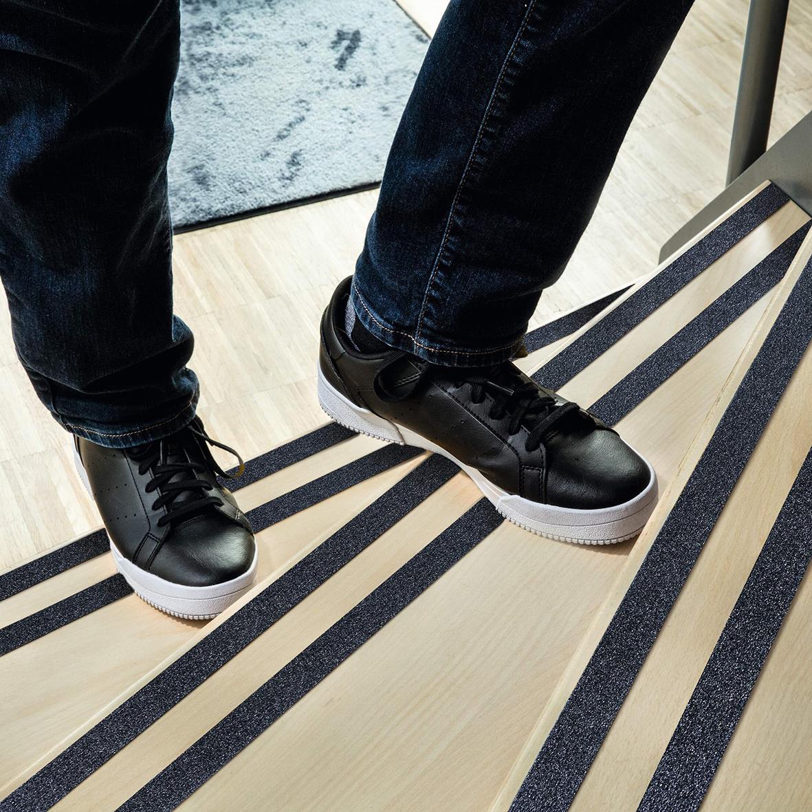 Person står på halkskyddstejp i svart och vitt, klädd i svarta jeans och sneakers på ett trägolv.