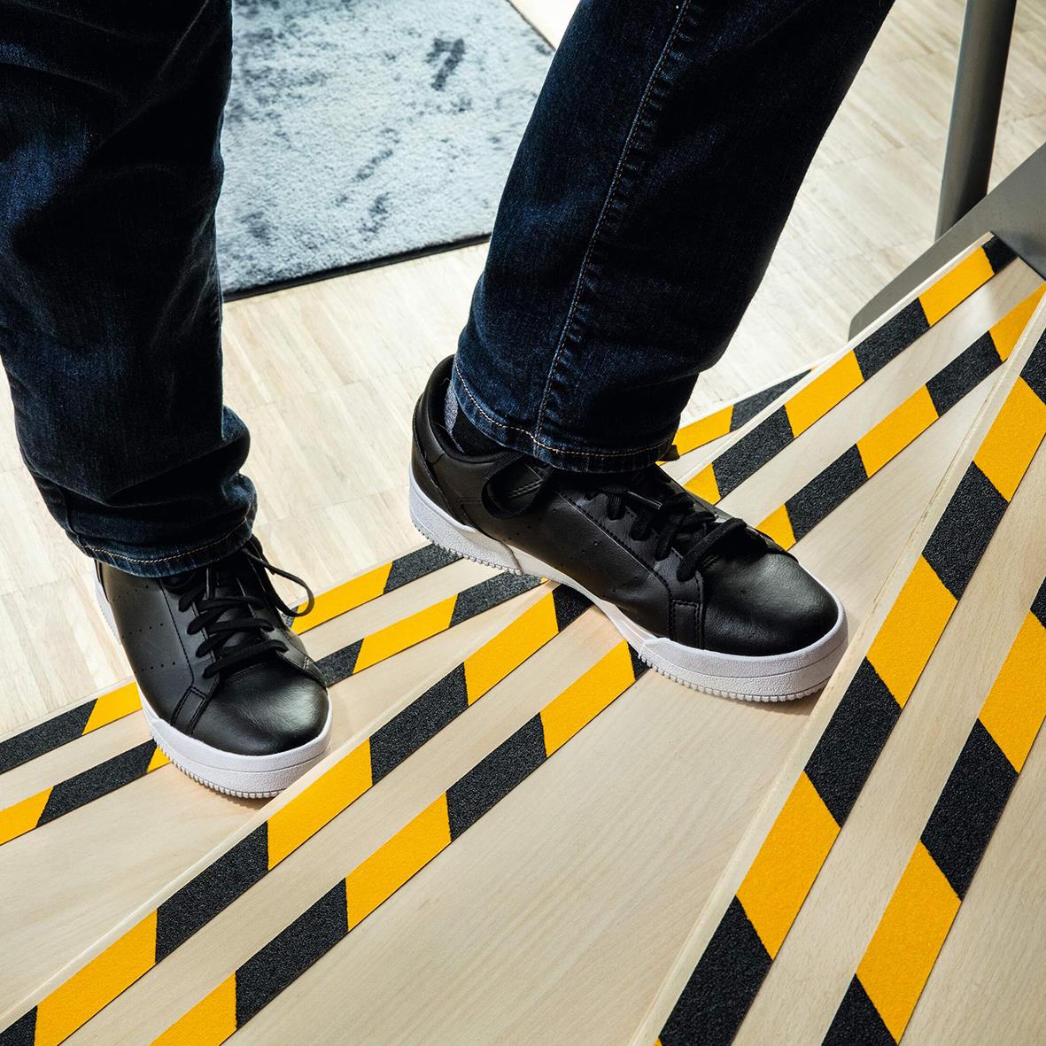Person står på halkskyddstejp i gul och svart på trappa, bär svarta sneakers och jeans