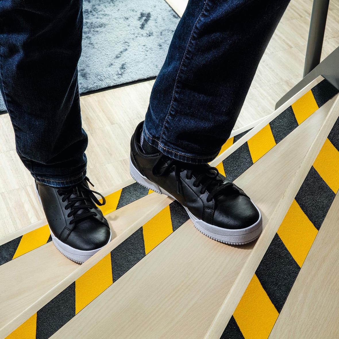 Person balanserar på en träbalk med halkskyddstejp i gul och svart färg, bär svarta sneakers, inomhusmiljö