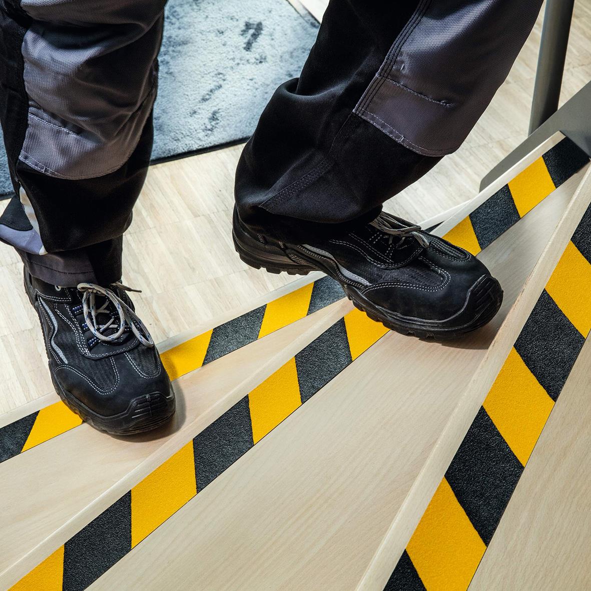 Person står på en trappa med halkskyddstejp i gul och svart för extra grepp, bär svarta skor och arbetskläder