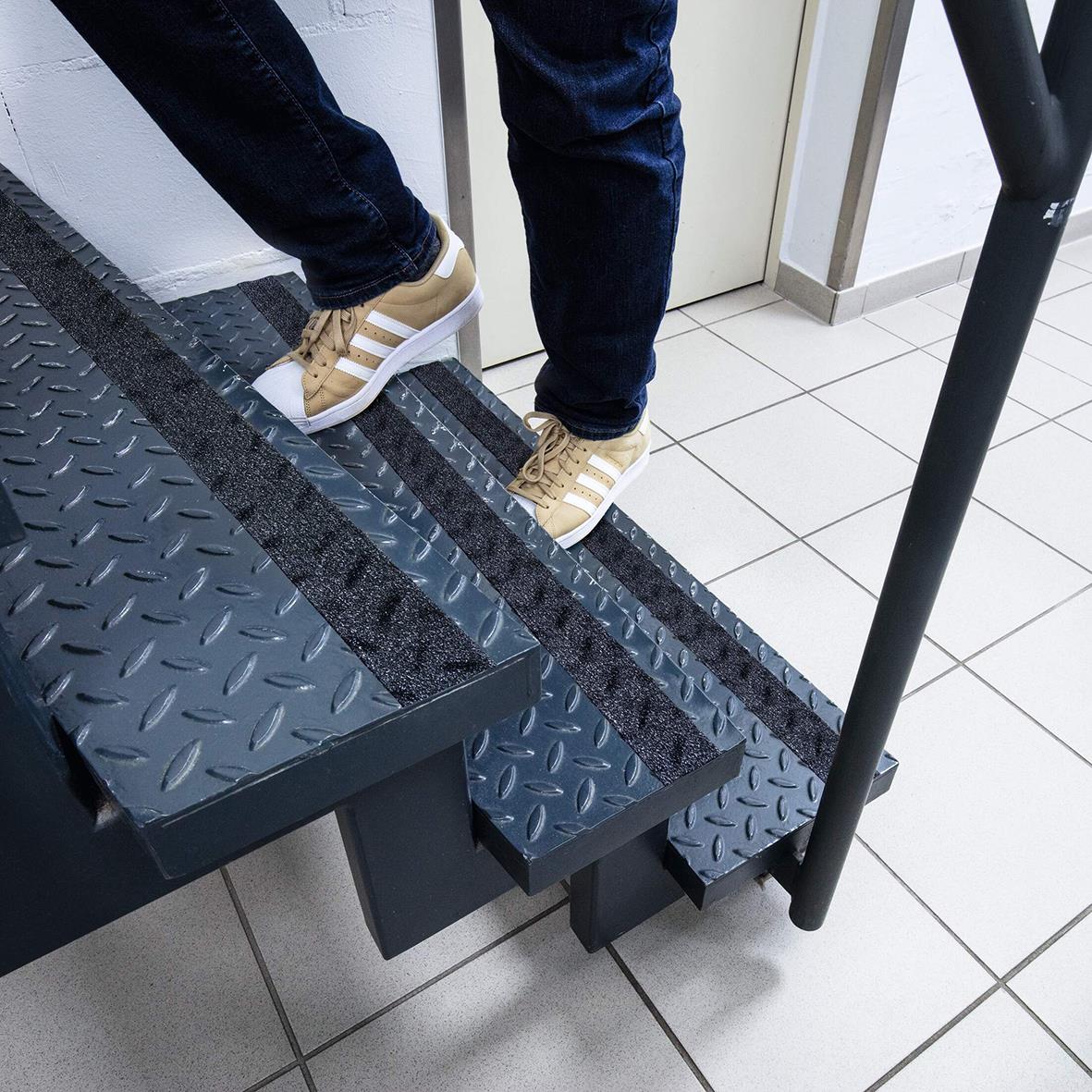 Person står på svart halkskyddstejp på trappa, klädd i jeans och sneakers i en inomhusmiljö