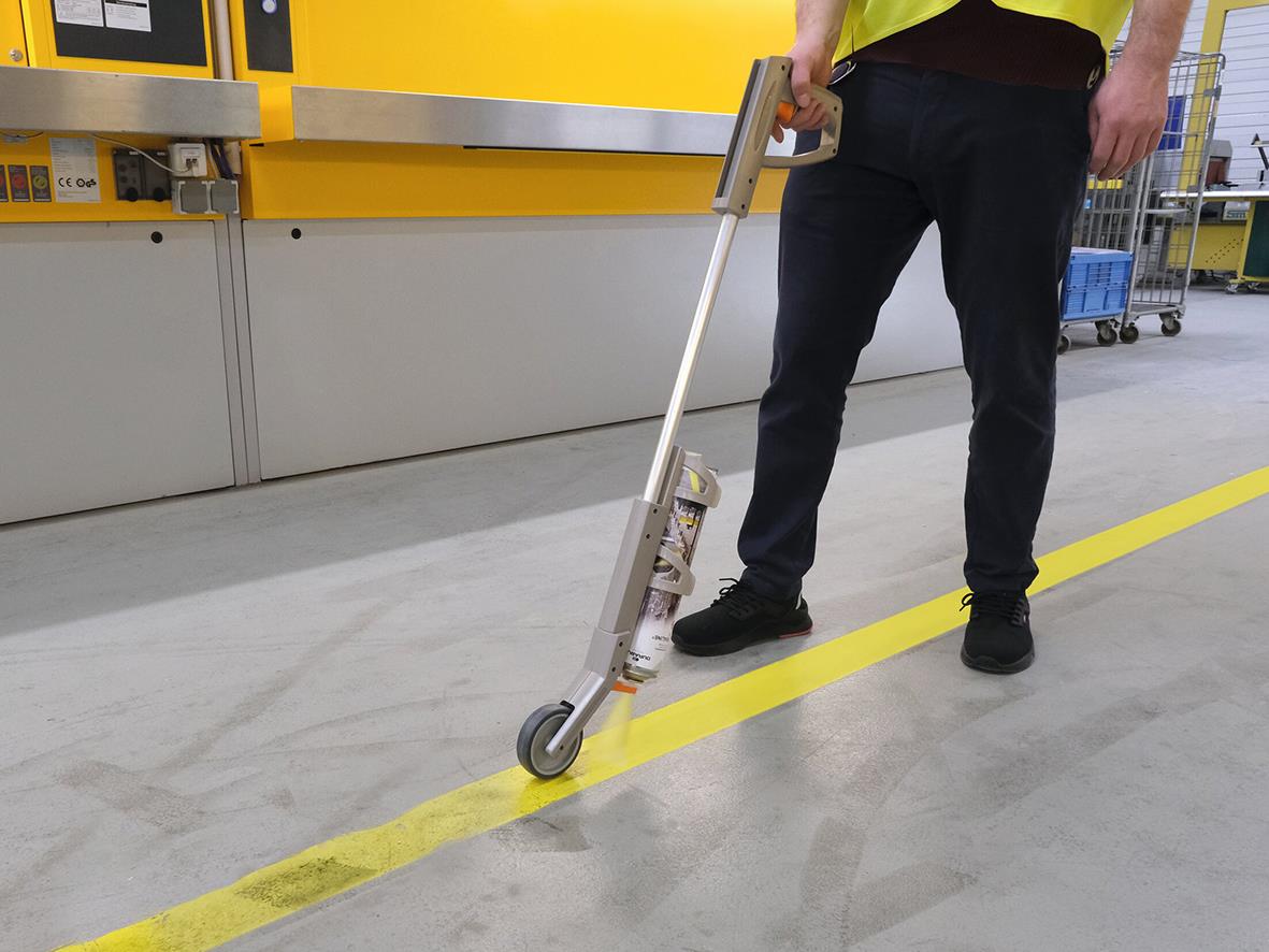 Person använder sprayhandtag för att måla en gul linje på golvet i ett lager eller fabrik