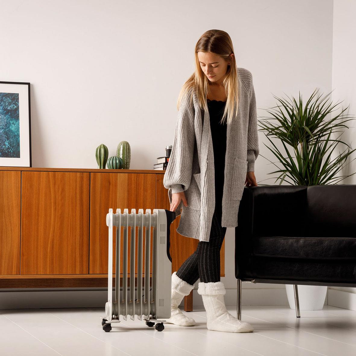 Kvinna med lång grå kofta och svarta byxor som flyttar en oljeradiator i ett modernt vardagsrum