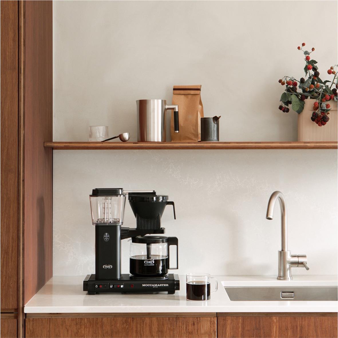 Kaffebryggare Moccamaster Optio Matt Svart på köksbänk med kaffekopp och växter i bakgrunden