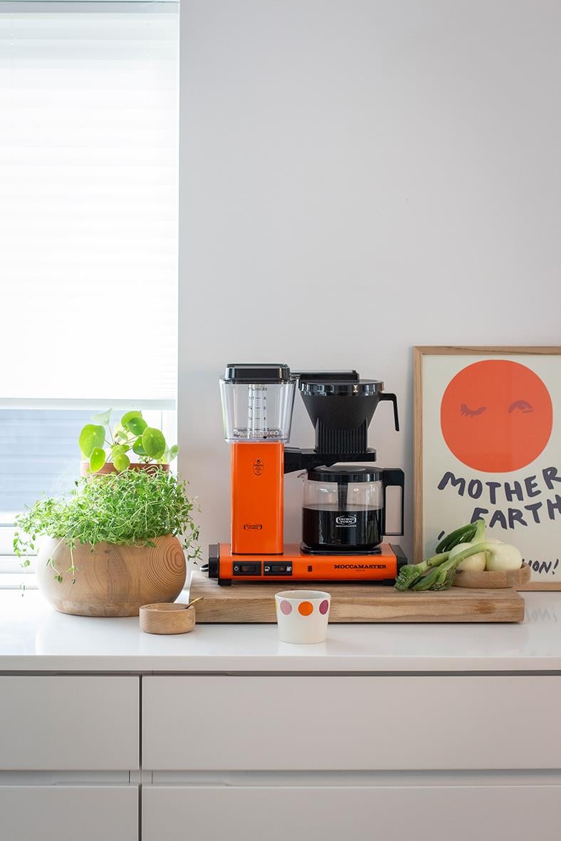 Kaffebryggare Moccamaster Optio Orange på köksbänk med växt och konstverk i bakgrunden