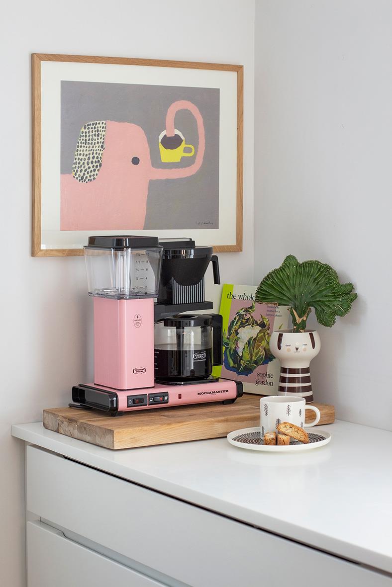 Pink Moccamaster kaffebryggare på köksbänk med kopp och kakor, konstverk och växt i bakgrunden