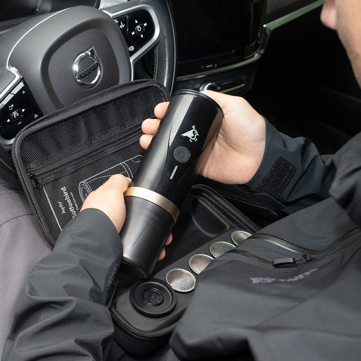 Person håller en batteridriven espressomaskin i bilen, med ett förvaringsfodral och kaffekapslar bredvid