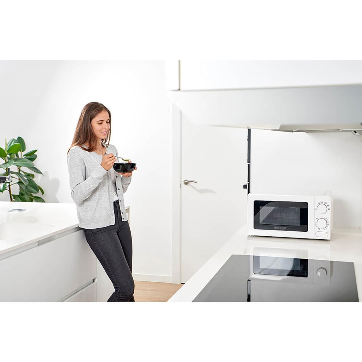 Kvinna äter mat framför mikrovågsugn i kök, vit Black+Decker mikrovågsugn med manuella vred