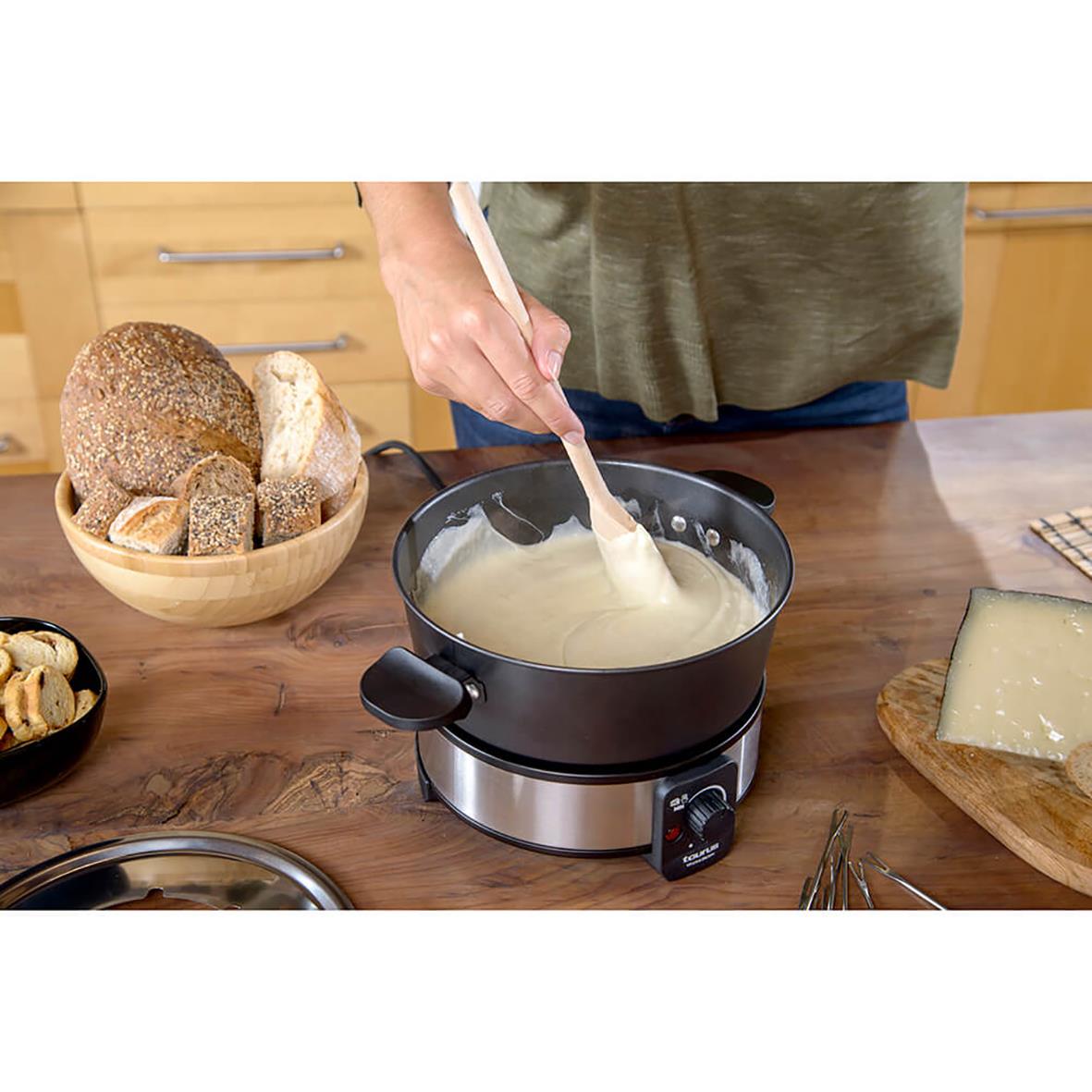 Person rör i en fonduegryta Taurus 1000W med ost, omgiven av bröd och ost på köksbordet