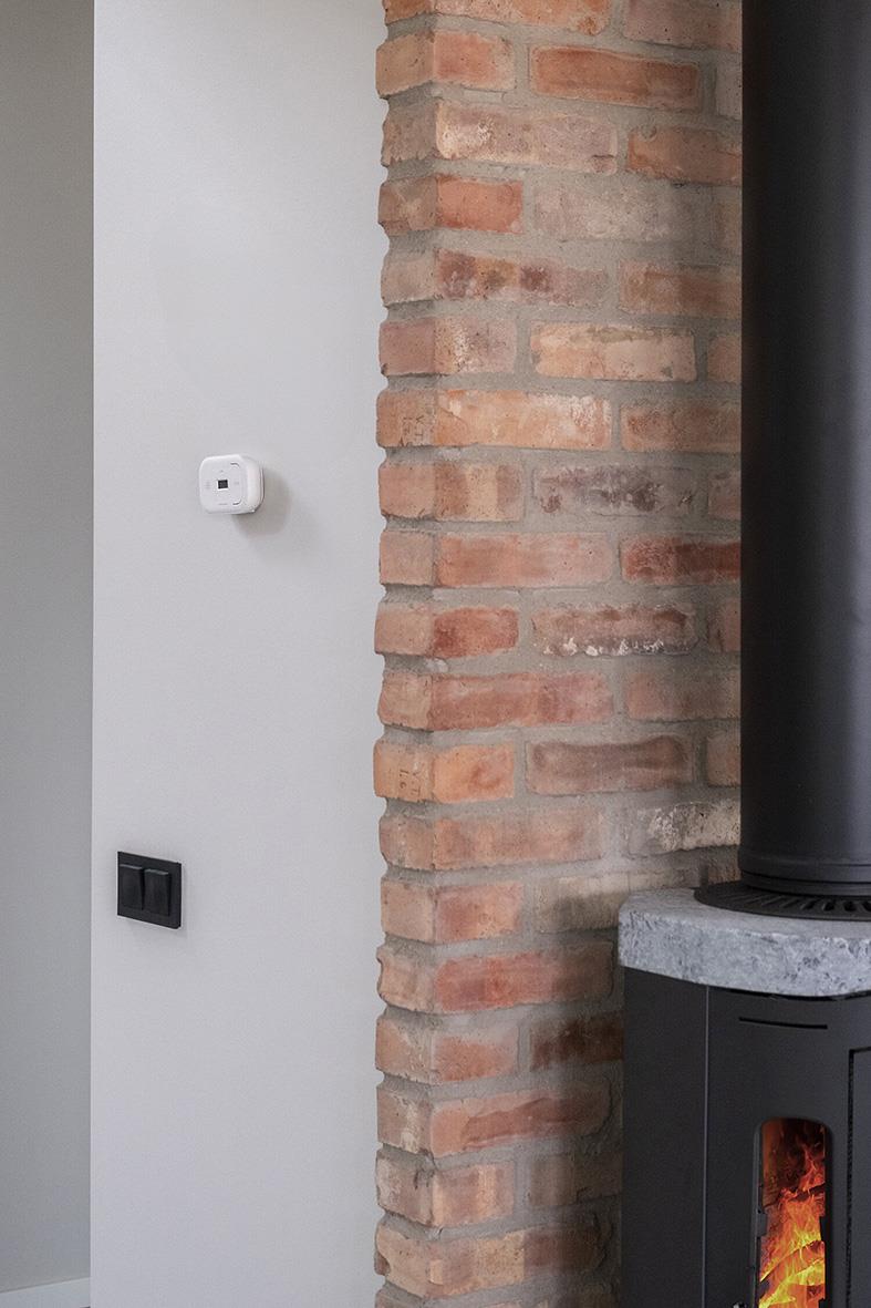 Kolmonoxidlarm Housegard CA108 med LCD-skärm monterad på vägg bredvid en tegelvägg och en eldstad