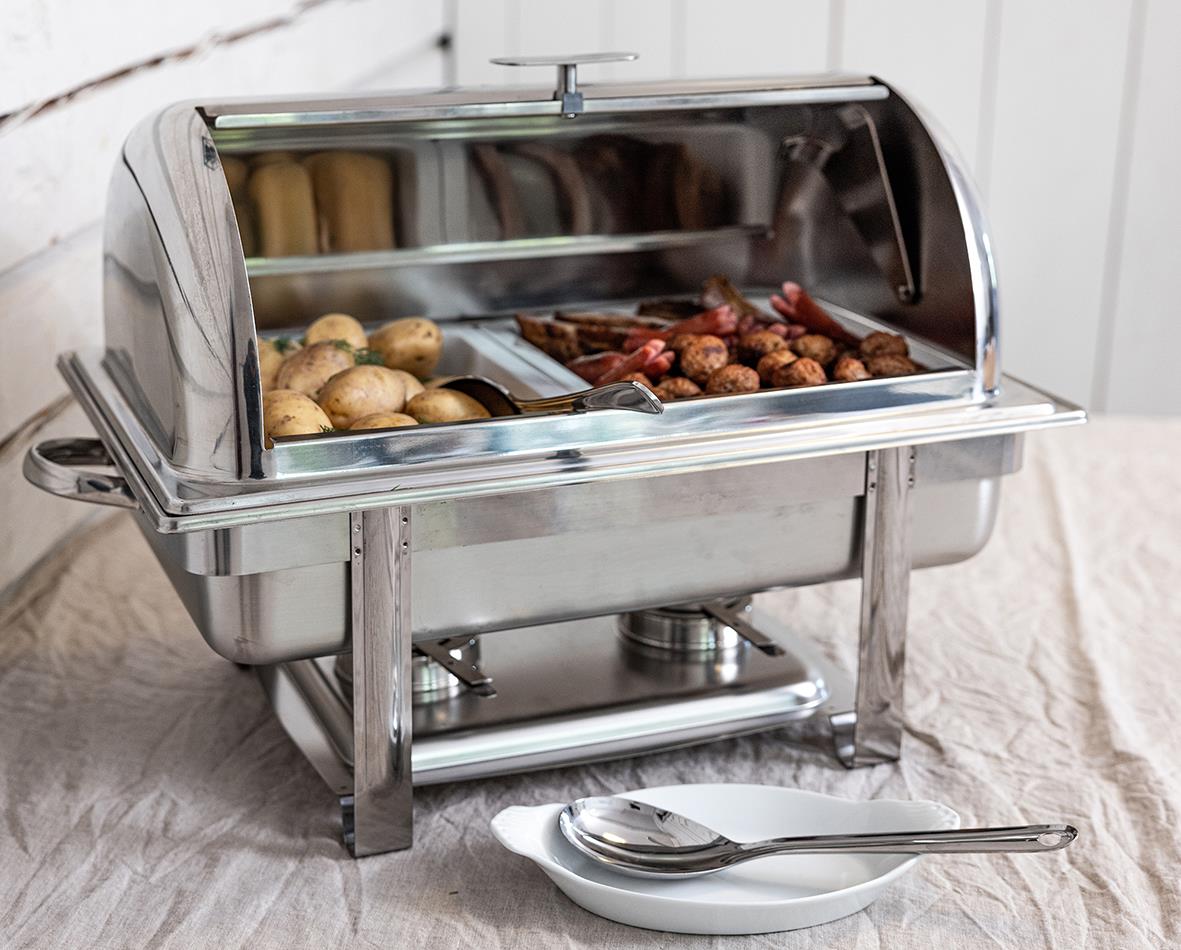 Stainless steel chafing dish med potatis och kött, placerad på ett bord med sked och serveringsfat
