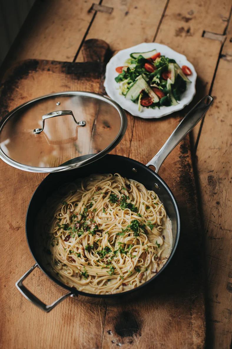 Rostfri traktörpanna med lock och pasta, bredvid en sallad på träbord