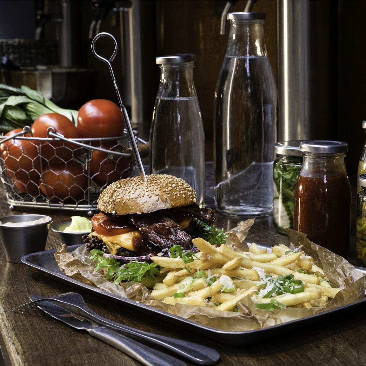 Hamburgare med pommes frites på svart tallrik, omgiven av ketchup, vattenflaskor och tomater i bakgrunden