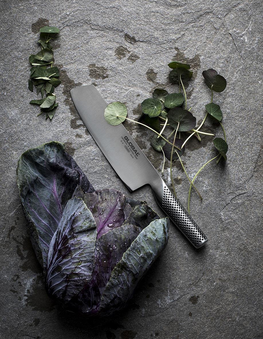 Grönsakshacka Global G-5 18cm med ett knivblad och gröna blad på en stenig yta
