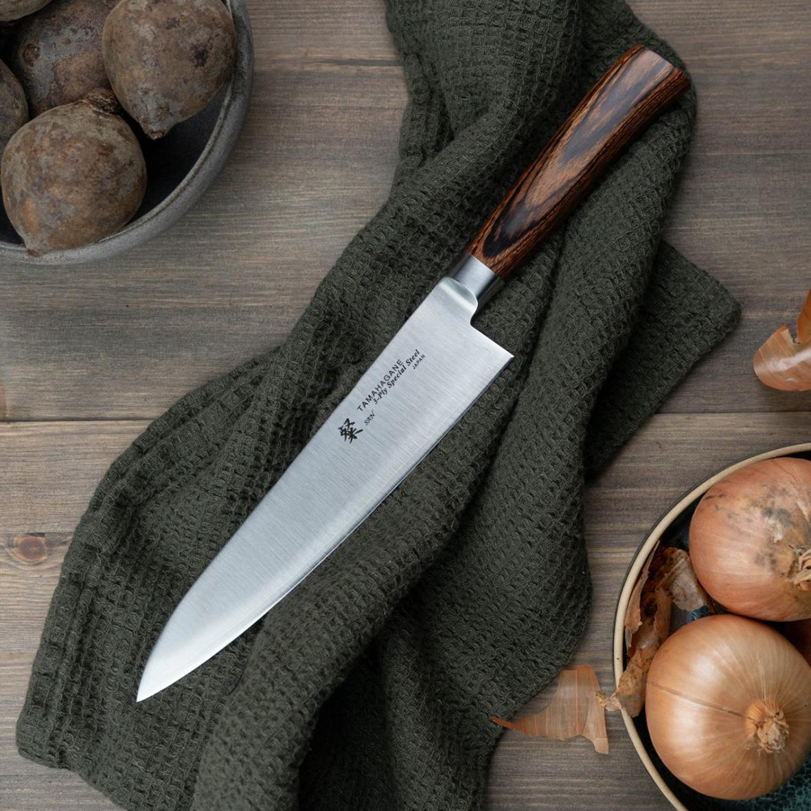 Kockkniv Tamahagane San 21cm med trähandtag vilande på grön duk omgiven av lök och rotfrukter