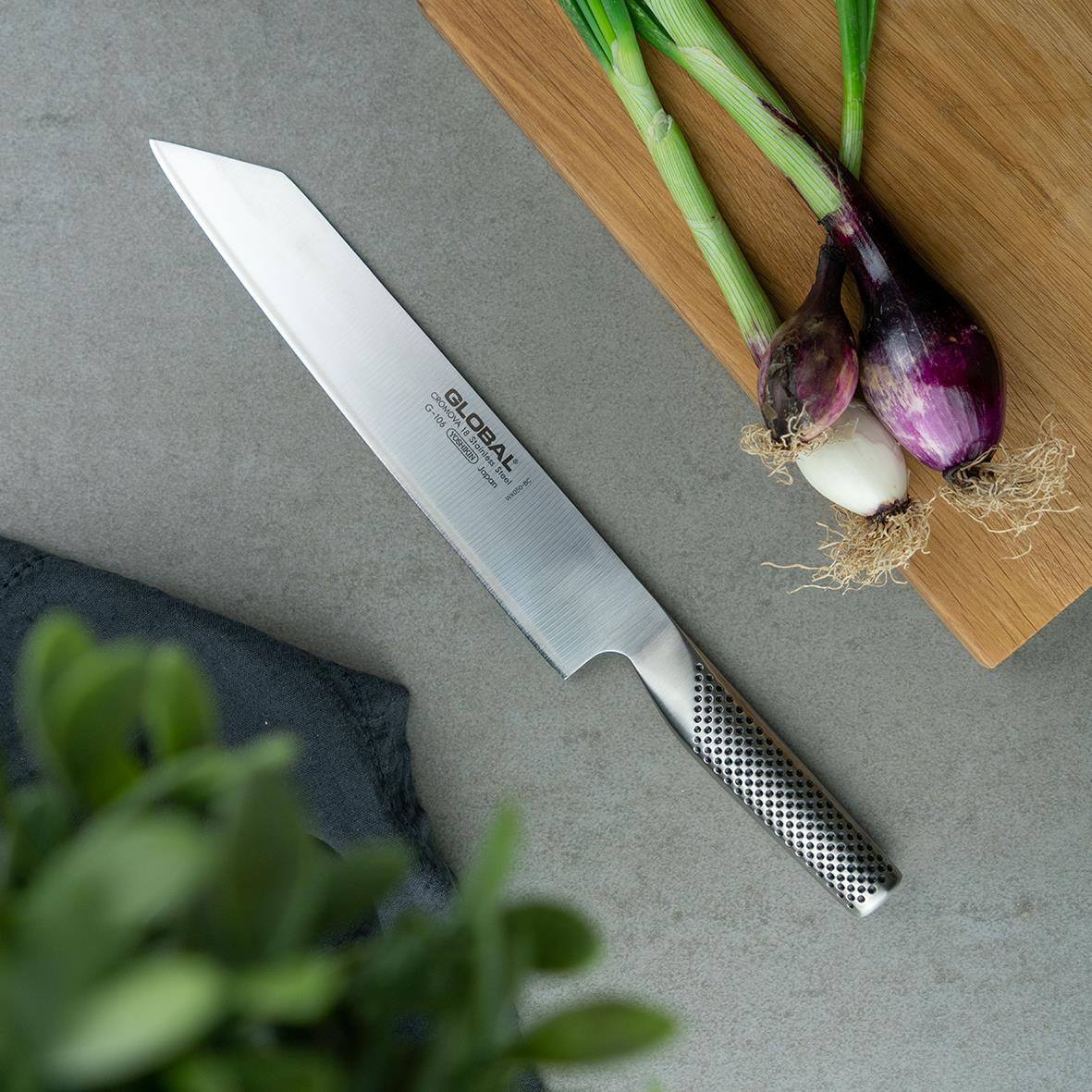 Kiritsuke Global G-106 24cm kniv med svart och vit handtag, placerad på en grå yta med gröna lökar och en träskiva i bakgrunden