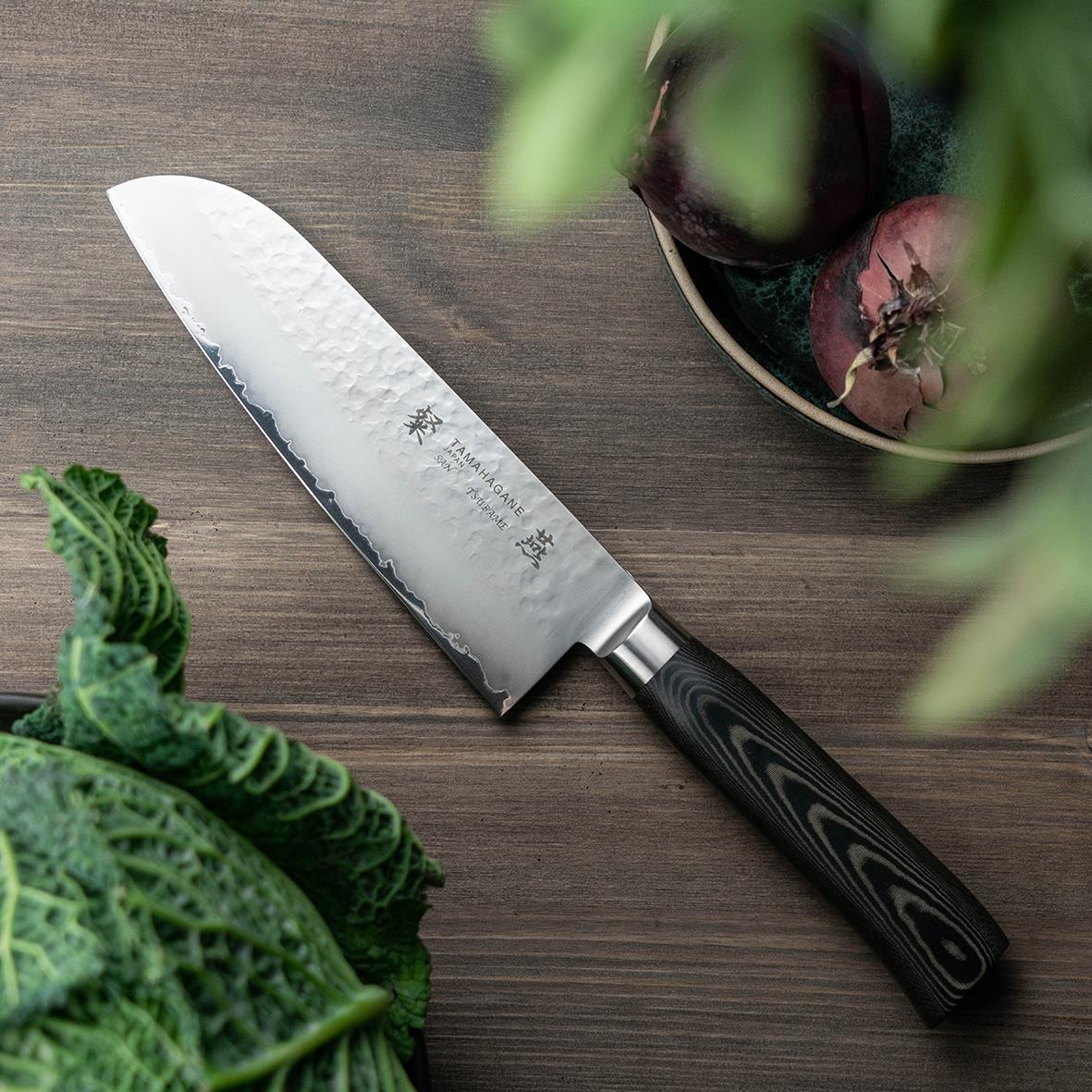 Santokukniv Tamahagane San Tsubame 16cm med svart skaft på träbord med gröna blad och växt