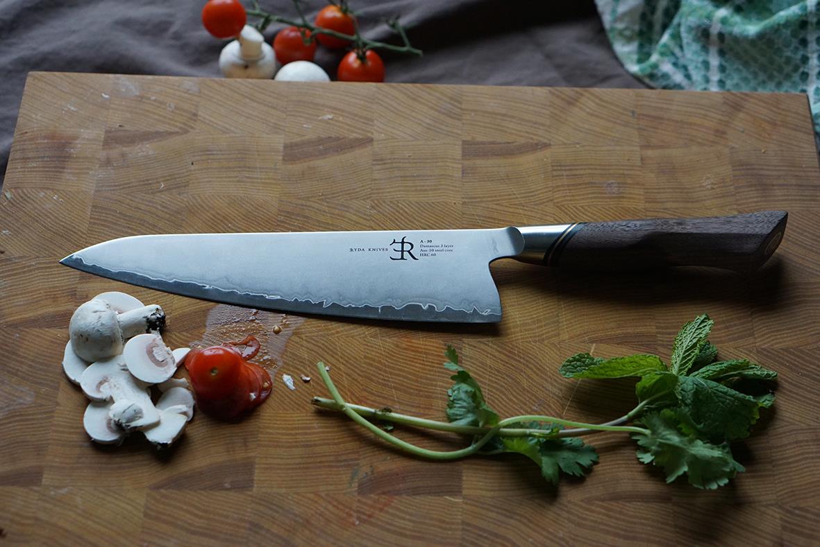Kockkniv Ryda Knives A-30 20cm placerad på träskärbräda med tomater, svamp och gröna örter