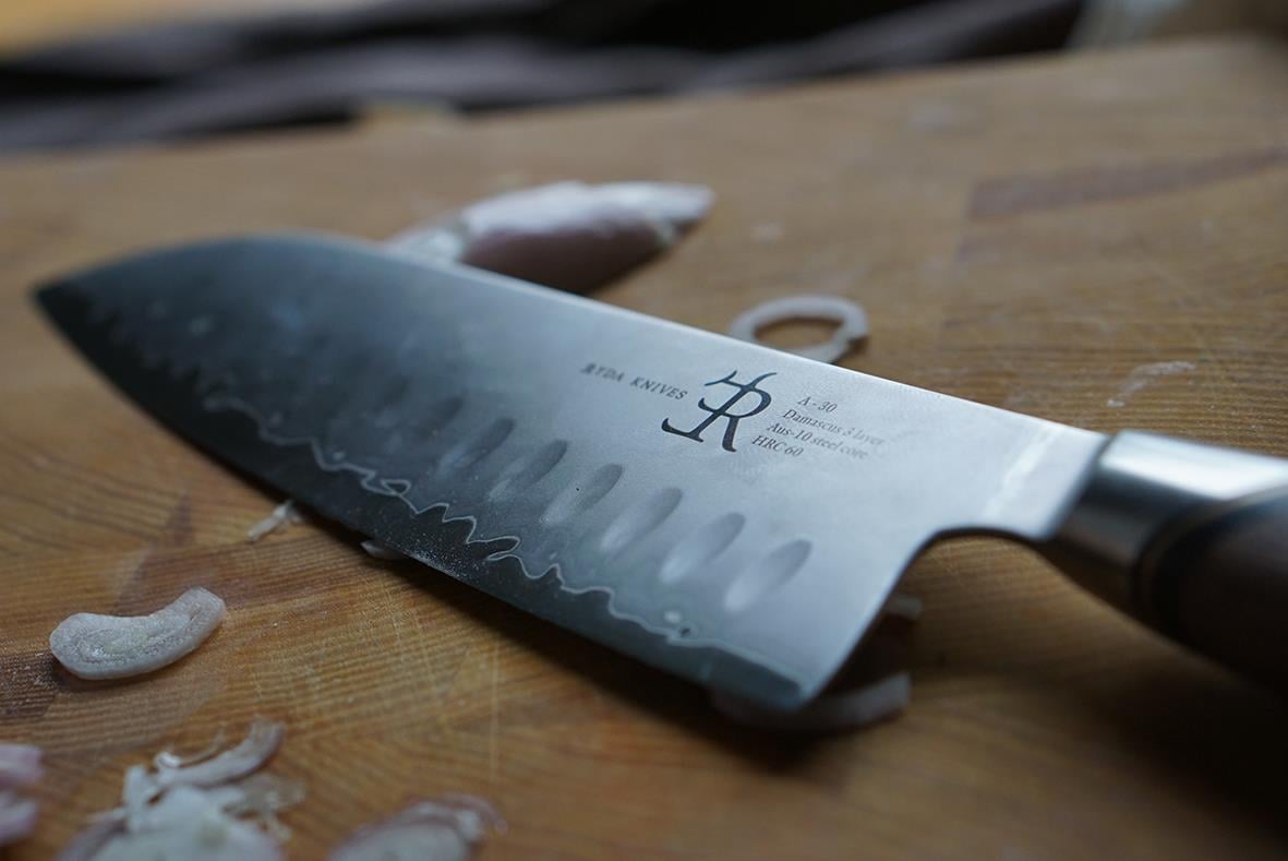 Santokukniv Ryda Knives A-30 18cm placerad på träbänk med skärbräda och skalor i bakgrunden
