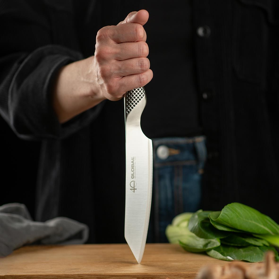 Person håller i en Kiritsuke Global 40-års Jubileum GS-110 kniv på 15 cm över ett träbord med gröna blad och en svart bakgrund