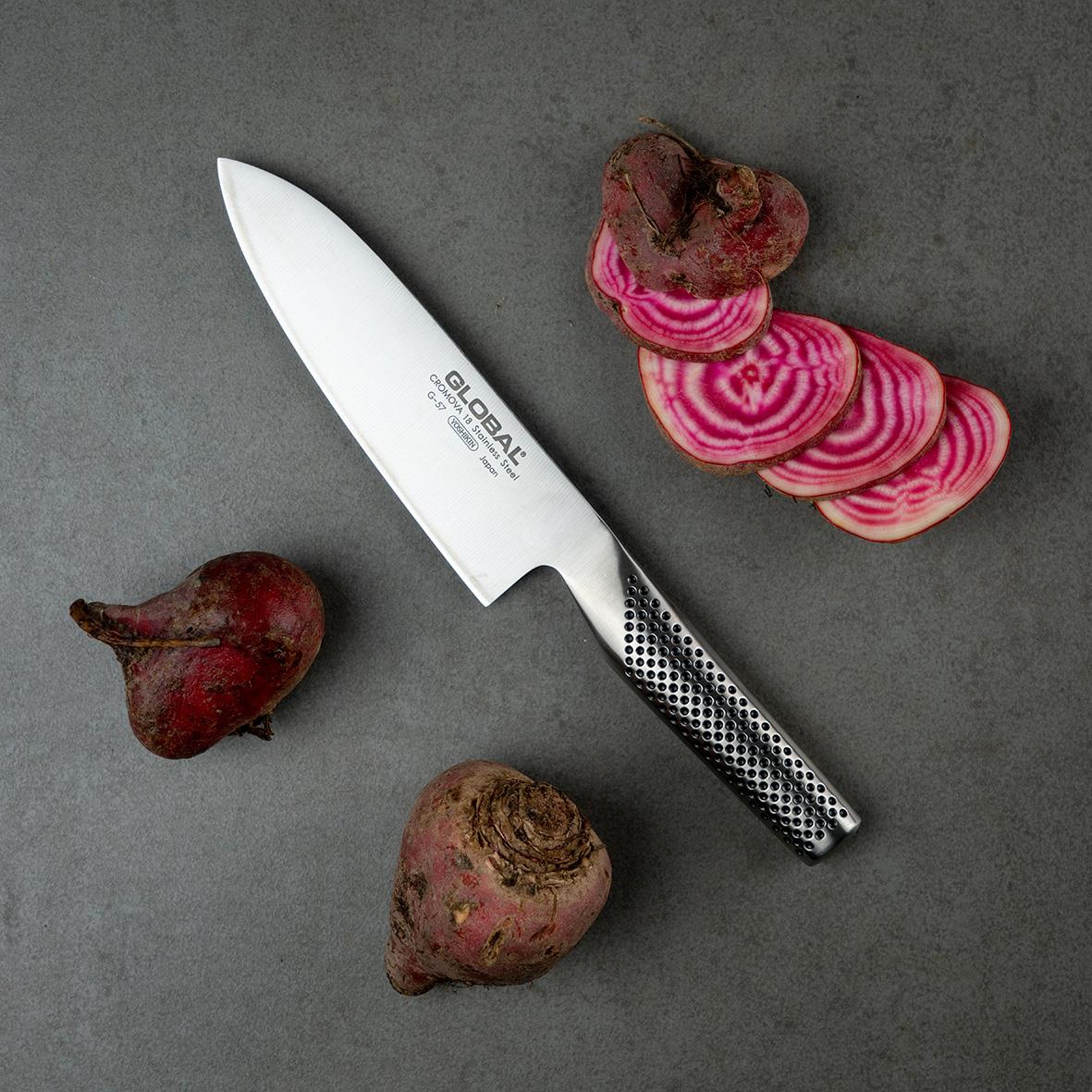Santokukniv Global G-57 16cm med svart och vitt mönstrad handtag, omgiven av röda och rosa rotfrukter på grå bakgrund
