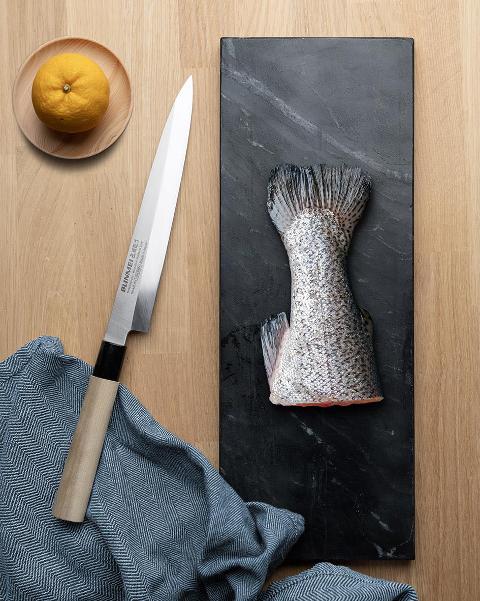 Sashimikniv Bunmei Yanagi på en svart stenplatta med en citron, kökshandduk och ett köksknivshandtag
