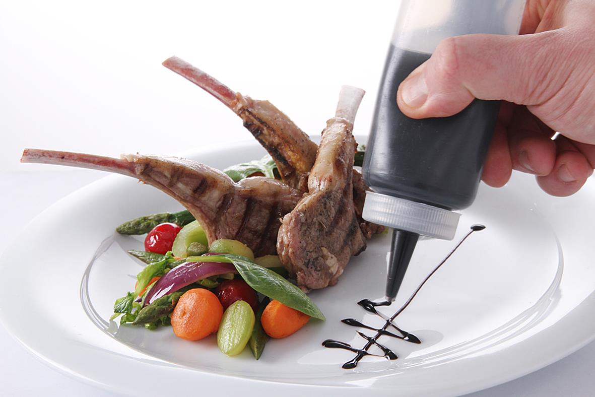 Person dekorerar en tallrik med kött och grönsaker med svart sås i en sprits.