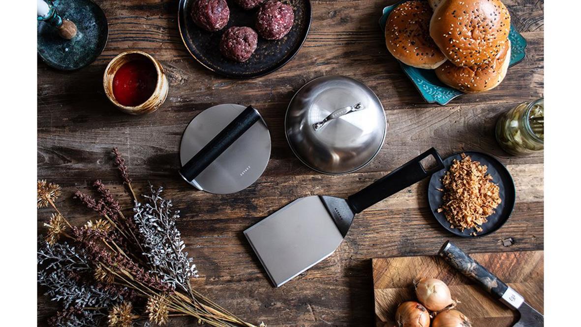 Smashburgerset med stekspade, press och cloche på träbord med bakverk och kryddor