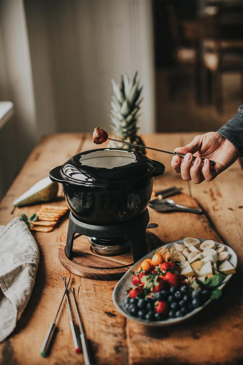 Fondueset i gjutjärn med rostfria gafflar på träbord, omgiven av frukt och tillbehör