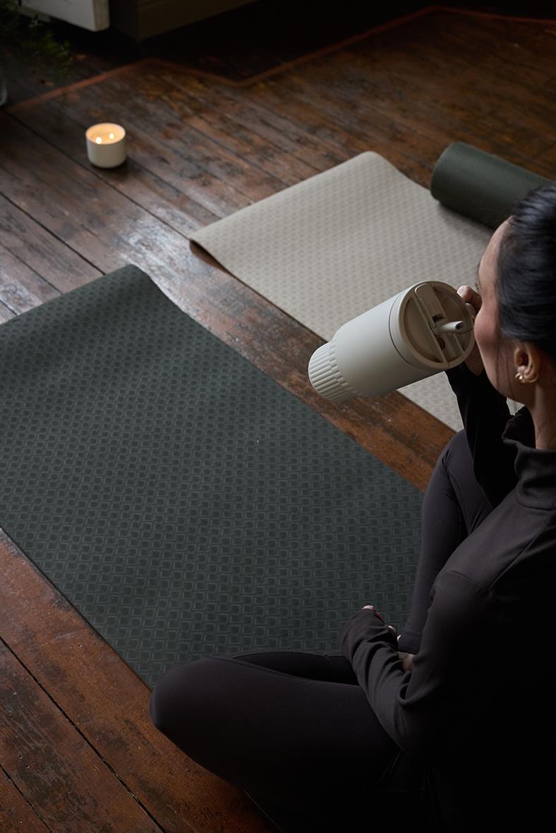 Kvinna dricker ur termosmugg på yogamatta i ett rum med trägolv och tända ljus