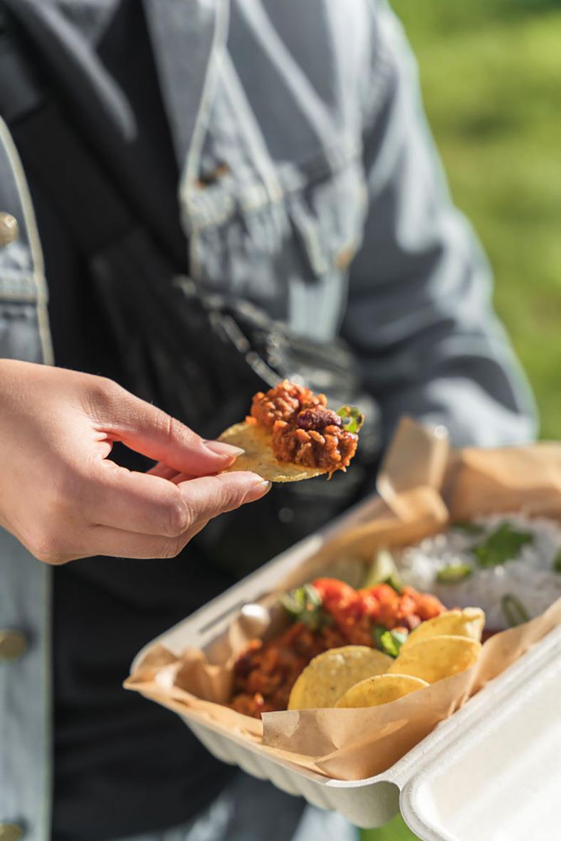 Person som håller i en bit mat framför en pappersform med mat, utomhusmiljö
