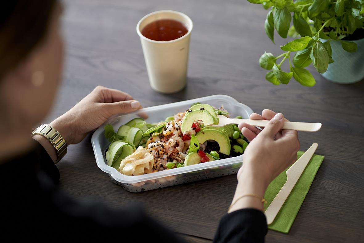Person äter sallad i plastlåda med grönt och avokado, på bord med dryck och växt
