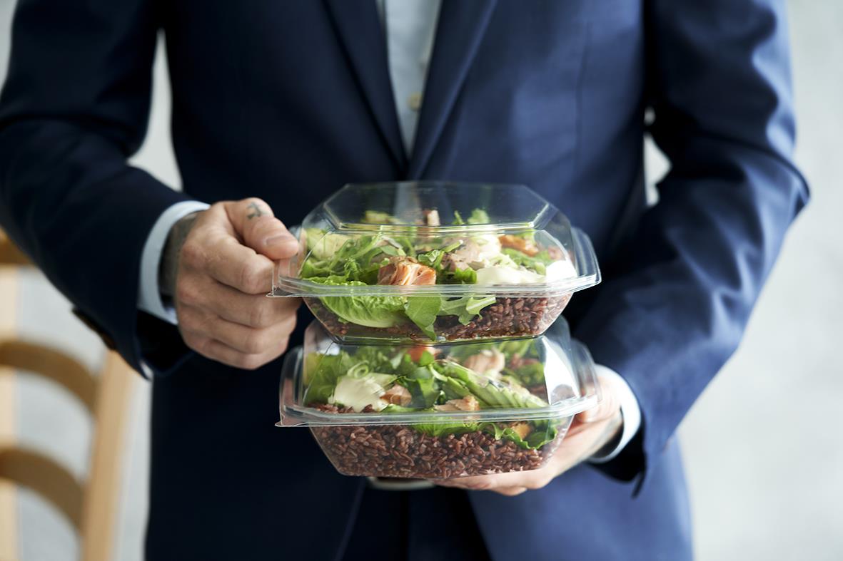 Person i kostym håller två plastbehållare med sallad och mat, transparenta och rektangulära