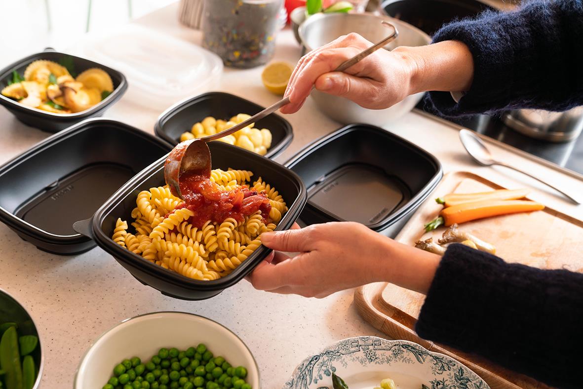 " som serverar mat i en svart plastlåda med lock, omgiven av andra matlådor och mat på ett köksbord"
