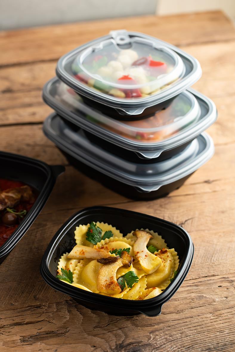 Flera svarta plastlådor med lock, en fylld med pasta och grönsaker, på träbord