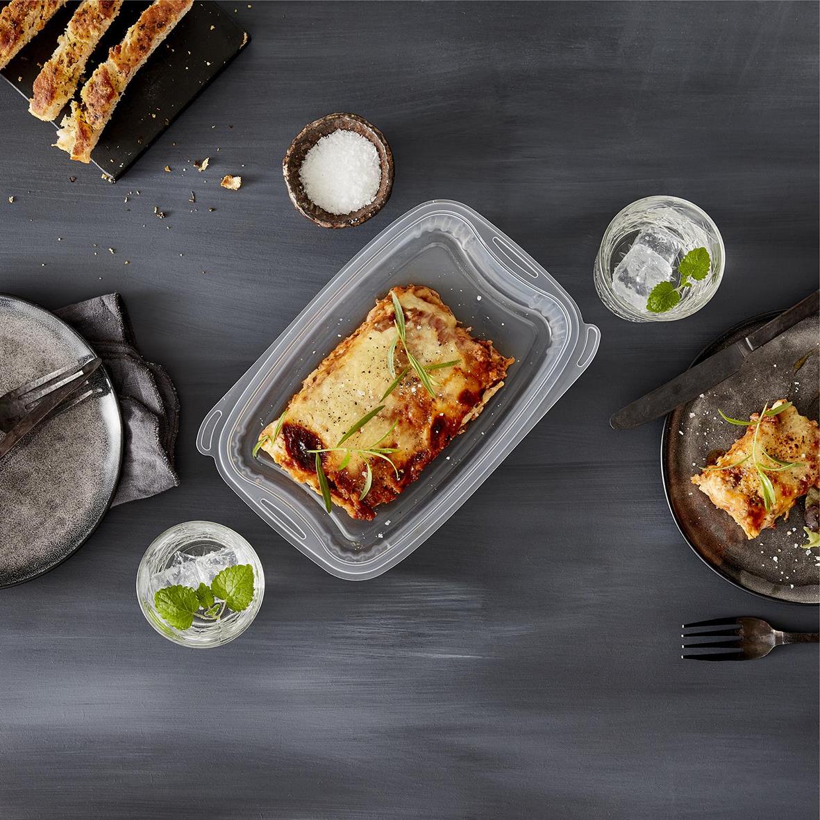 Plastform Bistro PP klar matlåda med lasagne, omgiven av salt, vatten och bröd på en mörk bakgrund