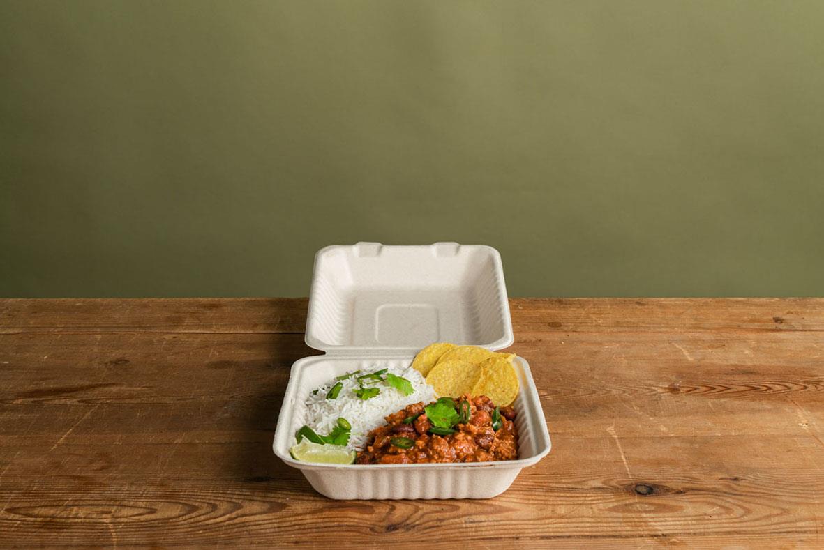 Hämtbox av bioplast med lock, innehållande ris, köttfärs och tillbehör, på träbord mot grön vägg