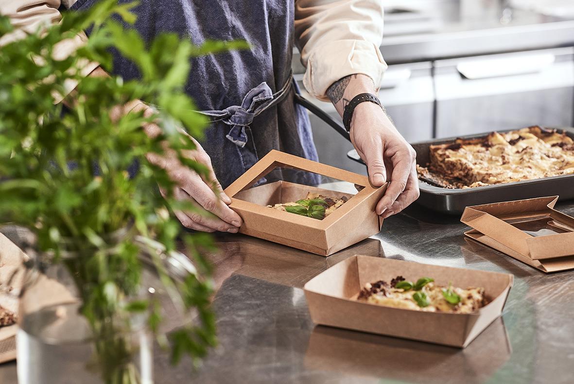 Person placerar mat i en låda med fönster, omgiven av mat och köksutrustning